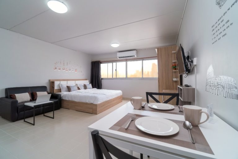 A well-furnished studio apartment in Dubai, featuring a cozy bed with white linens, a light gray sofa, and decorative elements. The living area includes a small black coffee table and a colorful rug, while the kitchenette is equipped with modern appliances, including a microwave and a refrigerator. The walls are adorned with framed art, and there are stylish lamps providing ambient lighting. This setup showcases an inviting space ideal for first-time residents on a budget.