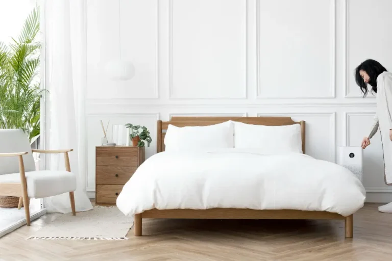 A woman stands in front of a bed with a white bedspread, smiling and dressed casually in a cozy bedroom setting.
